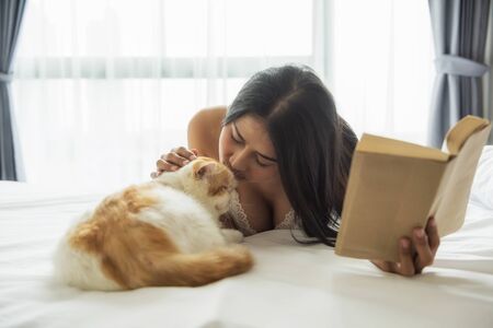 Beautiful attractive Asian tan woman with white pajama read book and kiss yellow exotic shorthair brown cat on bed in modern bedroom. Long hair sexy girl play with cute pet in morning.の写真素材