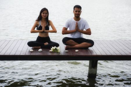 Healthy young Asian woman and man couple perform lotus yoga meditation hand pose on wood jetty with white rocks and Frangipani flowers above lake. Indian beard guy and Chinese girl.の写真素材