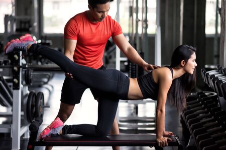 Portrait of personal trainer coaching beauty Asian woman to stretch back and leg in fitness sport gym. Happy girl working out  or yoga on chair for bodybuilding and healthy lifestyle.の写真素材