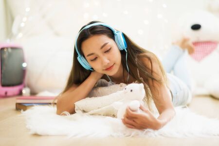 Beautiful attractive Asian girl look at bear doll, listen to online music, and think about her boyfriend in bedroom at winter with copy space for text. In love and miss conept.の写真素材