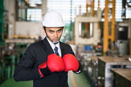 Asian businessman with safety helmet and red Boxing gloves ready for corporate battle. Man in suit shirt and tie holding combat gloves together. manufacture factory of relentless struggle and success.の写真素材