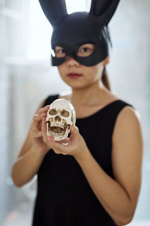 Asian girl with black bunny ear mask hold mock up skull with light from window. Female model for 2019 Halloween party concept with copy space for text.の写真素材