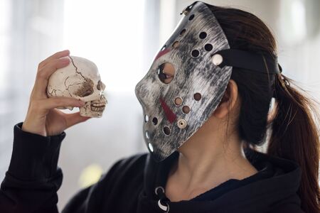 Asian woman with Jason mask and black hoodie dress look at mock up skull with light from window. Female model for 2019 Halloween party concept with copy space for text.の写真素材