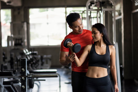 Portrait of personal fitness trainer or boyfriend coaching dumbbell exercise to sexy woman in sport gym. Happy couple working out for bodybuilding and healthy lifestyle.の写真素材