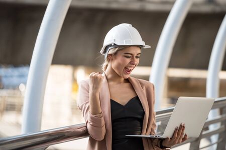 cheerful 20s American engineer woman with helmet and coat working on laptop while standing at urban city. Beautiful female worker celebrate construction  project or bidding complete from internet.の写真素材