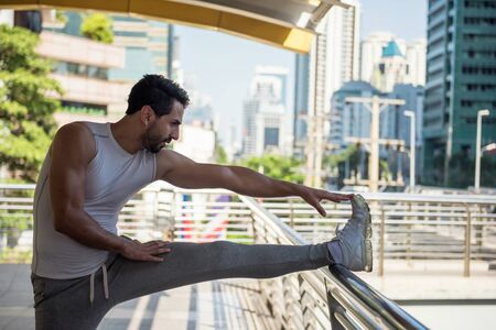 Athlete sporty Arab handsome beard man stretching foot and leg on skywalk  handrail before jogging or running with modern city building background. Sport and healthy lifestyleの写真素材