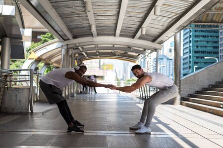 Young American male friends standing on city skywalk and holding hands to stretching before workout. urban skyline in the background. Sport and healthy lifestyle in town.の写真素材