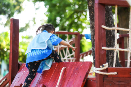 Kindergarten child girl climb up from wooden slope of kid playground with green foliage bokeh...