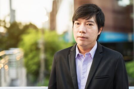 Portrait of Korean Businessman look at camera in modern city at sunset. Happy Asian entrepreneur, 30s to 40s at Bangkok business area and blurred modern building and space for text.の写真素材