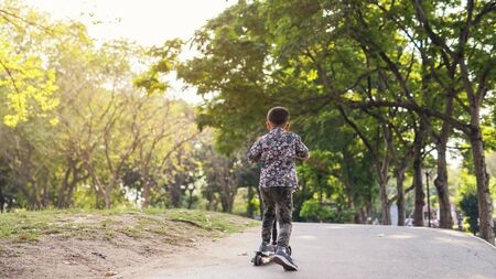 Boy rides uphill of the park by manual scooter with greenery trees and sunset background. Healthy sport lifestyle.の写真素材