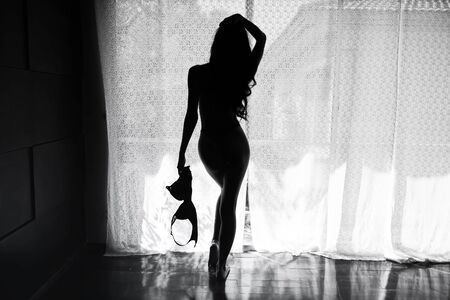 rear nude photo of beautiful Asian woman hold bra with sunlight via curtain. Silhouette sexy girl portrait, half nake, with black and white process.の写真素材