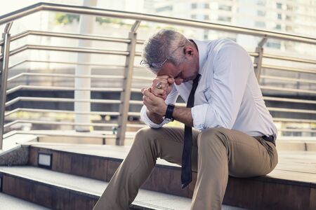Anxious senior businessman sitting in steps. Stressed gray hair businessman headache and look down with depress feeling. Business problems, Fired, Unemployed, bankrupt due to covid-19.の写真素材