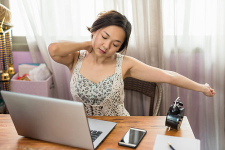 Beautiful Asian businesswoman in house stretching arm and massaging neck after hard working. Work...