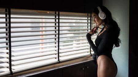 Alone Asian woman listen stream music by headphone on smartphone near window and miss her boyfriend. Social distancing, quarantine, stay at home to prevent COVID-19 or coronavirus.の写真素材