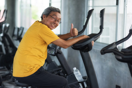 Happy senior aged man with gray hair exercising on cycling machine and thumb up in fitness gym. Old mature fit people biking cardio. Bodybuilding and elderly Healthy lifestyle.の写真素材