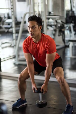 Asian muscular strong handsome man working out biceps by heavy dumbbell in fitness gym with sport exercise equipment background. Bodybuilding and healthy lifestyle concept.の写真素材
