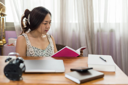 Asian Chinese woman sit and read book on table. Cute girl stay home to prevent coronavirus or...