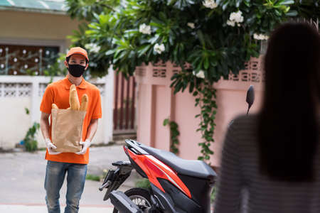 Asian delivered man with face mask and gloves in orange uniform hold bag of bread food deliver to female costumer in front of home. Postman and express grocery delivery by motorcycle during covid19.の写真素材