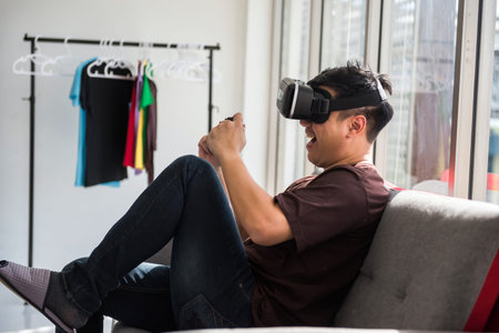 Portrait of excited Asian man on sofa play online game by VR glasses and wireless joystick in house....