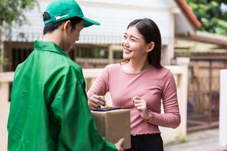 Beautiful smiling Asian client woman signing receive signature on smartphone after accepting a delivery of boxes from deliveryman in green jacket uniform.の写真素材