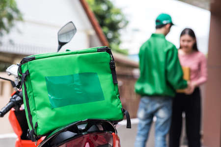 deliver motorcycle with isothermal food green case box  parking in front of house or home with blur courier delivery man and client. Deliver food at office building. Express delivering food In Cityの写真素材