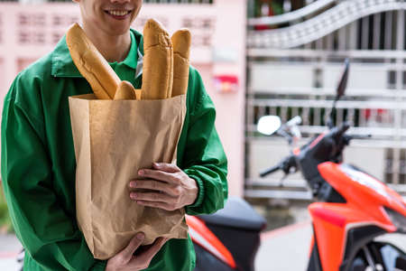 Smiling delivery man hold baguette bread in bakery paper bag to deliver customer with motorbike background. New normal of delivering business during covid19 or coronavirus pandemic concept.の写真素材