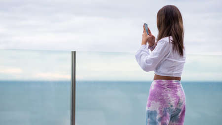 Young tan Asian woman photo sea coast view by smartphone on roof top of highrise hotel. Attractive girl holiday vacation to beach in summer with copy space for text.の写真素材