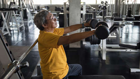 Old senior Asian man lifting and stretching dumbbells  by both hands in fitness gym with sport equipment bacground. Biceps exercise, bodybuiliding and heatlhy lifestyle for elderly pensioner.の写真素材