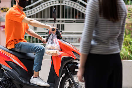 Food deliveryman with face mask in orange uniform wait customer on motorbike. Customer late to pick up online bakery. COVID-19 pandemic. New normal to use smartphone app to order.の写真素材