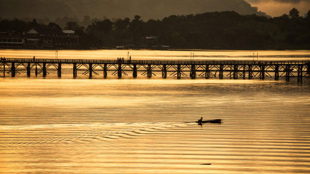 Silhouette fishermen sailing boat near Mon wooden or Attanusorn bridge at sunrise, Sangkhla Buri, Kanchanaburi, Thailand. village people fishing lifestyle in early morning.の写真素材