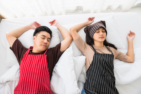 Tired couple with apron sleep or nap on bed by top view after cleaning home at bedroom. Housework on weekend of young exausted Asian husband and wife. End of house cleaningの写真素材