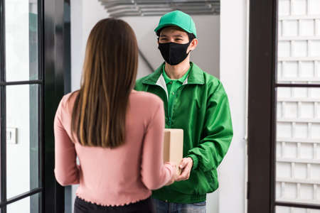 Rear of office woman receiving parcel from Asian courier deliveryman in uniform and face mask at door entrance. New normal to shopping by mobie phone application during covid19.の写真素材