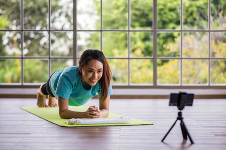 Young Asian athlete plank exercise on gym Mat. Girl blogger doing sports, looking at the camera and is live on smartphone at home.  online broadcast tutorial class.の写真素材