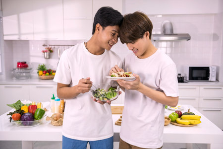 Happy LGBT Asian couple enjoy holding salad and sandwich to eat after cooking in kitchen. Lovely gay...