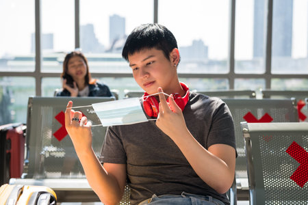 Handsome Asian young man wearing face mask at airport terminal seats. He wait for departure or...