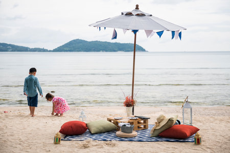 Beach picnic seat with champaign and food under parasol while kids, older brother and little sister, play sand in Phuket, Thailand. Family holiday vacation in summer.の写真素材