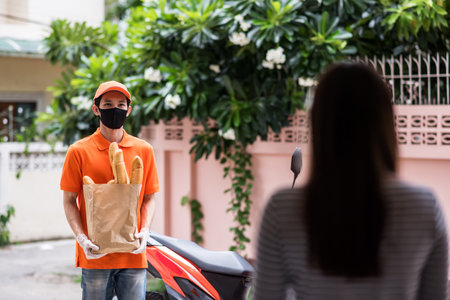 Asian courier man with face mask and gloves in orange uniform deliver bread to woman in front of home. express grocery delivery by motorbike during covid-19. New normal business.の写真素材