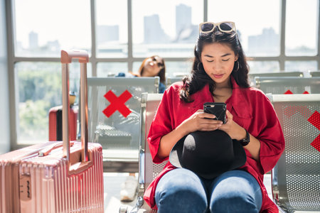beautiful Asian young woman waiting for flight departure, sitting in airport terminal lounge with phone in her hands. Social distancing for transportation travel in new normal of covid-19.の写真素材