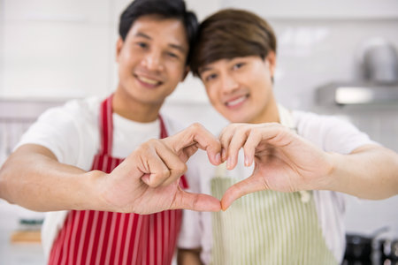 LGBT gay couple gesturing hands. Lovely homosexual Asian men make heart shape from fingers and palms. Same sex family celebrate valentine day in kitchen home.の写真素材