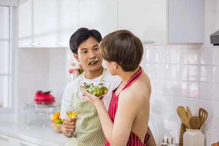 Sexy young topless LBGT man with red apron feeding salad to his boyfriend at kitchen after cooking to celebrate valentine's day. Lovely gay couple. Healthy eating lifestyle for same sex family.の写真素材