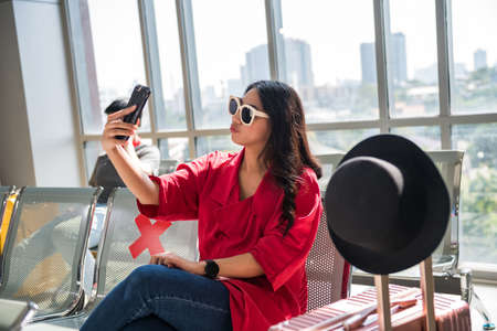 Young Asian beautiful woman selfie herself by smartphone at waiting seat zone with social distancing sign in airport terminal to wait for departure. Travel in new normal to prevent covid-19.の写真素材