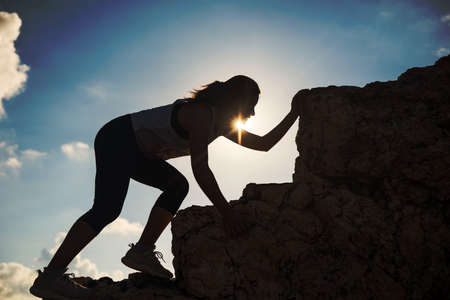 Silhouette hiking Asian young fit woman climb up to top of hill summit at sunset. Sporty female climber to mountain peak against sun rays. Bodybuilding by extreme sport.の写真素材