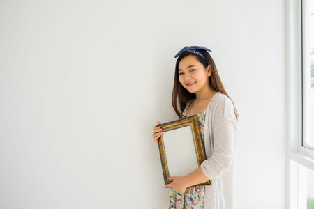 Portrait of young beautiful Asian woman hold gold picture frame to plan decoration on blank white wall of new house. Start new life by buying home. Real eatate and Property industry.の写真素材