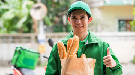 Smiling Asian deliveryman thumb up and hold baguette bread in bakery paper bag to deliver customer near motorbike. New normal of delivering business during covid-19 pandemic concept.の写真素材