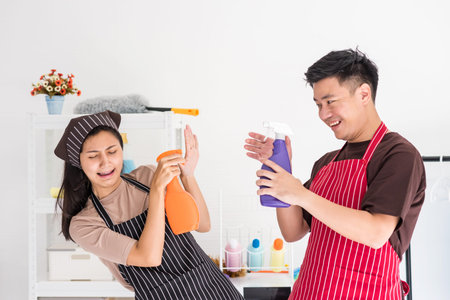 Asian couple having fun during cleaning house. Young husband spray water to his wife while pity girl...