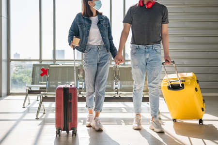 Traveller young Asian couple with face mask holding hands and wait for departure in airport terminal. Holiday maker or sweet vacation. New normal to travel and refrain from covid-19 pandemic.の写真素材