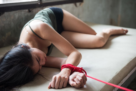 Sad young woman hostage lying on bed and tied by red rope in abandon house. Victim female kidnapped by robber. Sexual abuse or rape in dirty room with copy space for text.の写真素材