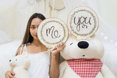 Portrait of young beautiful Asian woman hold me banner near big white bear with you banner at bedroom. Cute brunette girl play with her big fluffy doll at bed.の写真素材