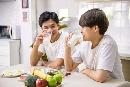 Happy Asain LGBT gay male couple drink milk, eat sandwich, salad for breakfast at home. They eye contact together with smiling face in dining room. Same sex family.の写真素材