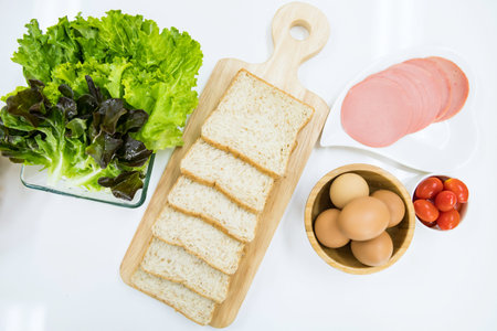 Top view of raw food ingredient material, wholewheat bread, boiled eggs, tomata, ham, lettuce vegatable preparation to make sandwich on white kitchen table. Healthy cooking and eating lifestyle.の写真素材